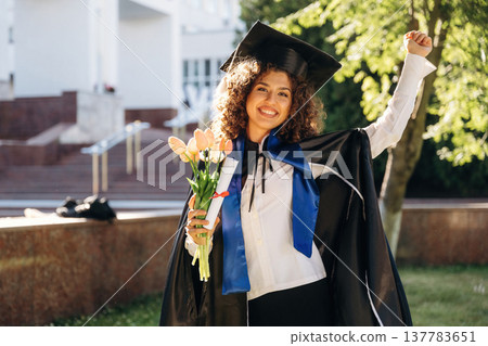 Flowers in hand. Cheerful graduate student in a gown is standing outdoors 137783651