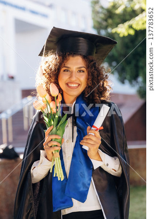 Flowers in hand. Cheerful graduate student in a gown is standing outdoors Flowers in hand. Cheerful graduate student in a gown is standing outdoors 137783652