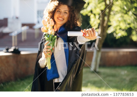 Flowers in hand. Cheerful graduate student in a gown is standing outdoors Flowers in hand. Cheerful graduate student in a gown is standing outdoors 137783653