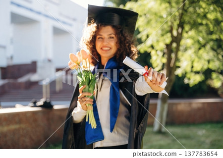 Flowers in hand. Cheerful graduate student in a gown is standing outdoors 137783654