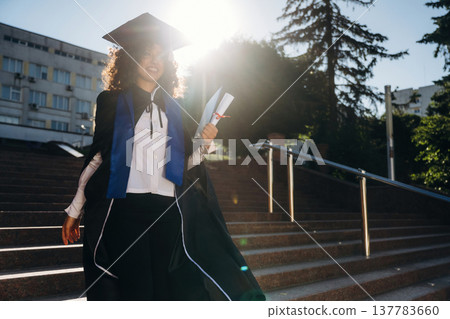 Stairs and university behind. Cheerful graduate student in a gown is standing outdoors Stairs and university behind. Cheerful graduate student in a gown is standing outdoors 137783660