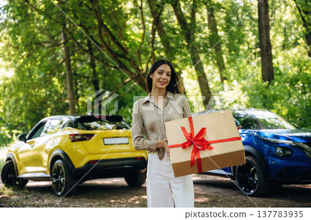 Present box in hands, standing. Young woman is in the forest with two cars 137783935