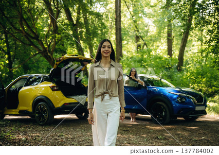 Posing and standing. Two women are near yellow and blue cars in the forest 137784002