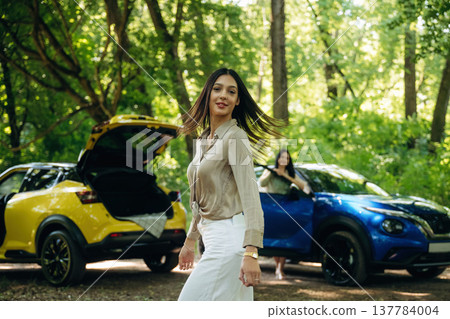 Posing and standing. Two women are near yellow and blue cars in the forest 137784004