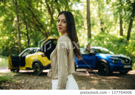 Posing and standing. Two women are near yellow and blue cars in the forest 137784006
