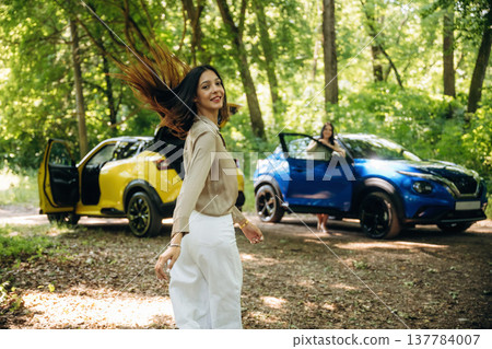 Posing and standing. Two women are near yellow and blue cars in the forest 137784007