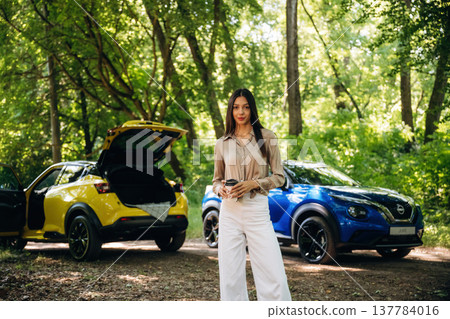 Holding cup of coffee in hands. Young woman is in the forest with two cars Holding cup of coffee in hands. Young woman is in the forest with two cars 137784016