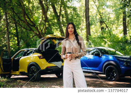 Holding cup of coffee in hands. Young woman is in the forest with two cars Holding cup of coffee in hands. Young woman is in the forest with two cars 137784017