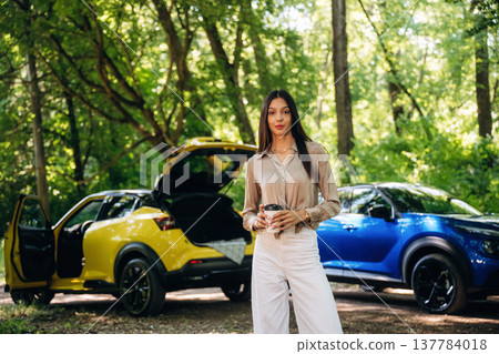 Holding cup of coffee in hands. Young woman is in the forest with two cars Holding cup of coffee in hands. Young woman is in the forest with two cars 137784018
