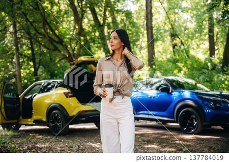Holding cup of coffee in hands. Young woman is in the forest with two cars 137784019