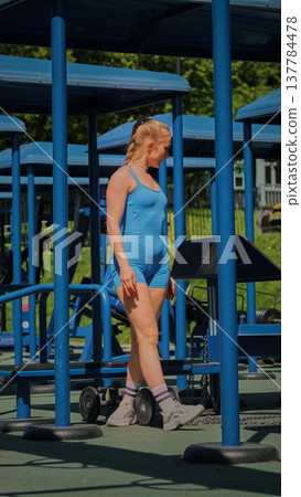 A woman in a blue sports outfit is standing next to gym equipment in an outdoor gym, preparing for her workout. This image highlights the importance of outdoor exercise for maintaining physical A woman in a blue sports outfit is standing next to gym equipment in an outdoor gym, preparing for her workout. This image highlights the importance of outdoor exercise for maintaining physical 137784478