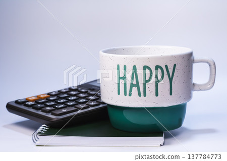 Ceramic coffee mug with Happy text placed beside calculator on spiral notebook isolated on white background. Business finance, accounting and productive workplace concept. 137784773