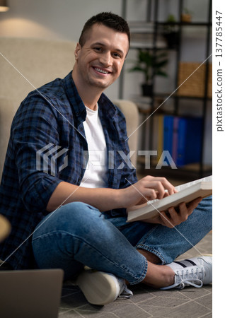 Positive man sitting on floor reading book in modern living space Positive man sitting on floor reading book in modern living space 137785447