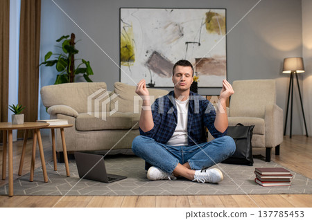 Calm man meditating with closed eyes sitting in lotus position on floor in living room Calm man meditating with closed eyes sitting in lotus position on floor in living room 137785453