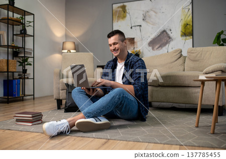 Joyful man using laptop on floor in modern living space with sofa and picture Joyful man using laptop on floor in modern living space with sofa and picture 137785455