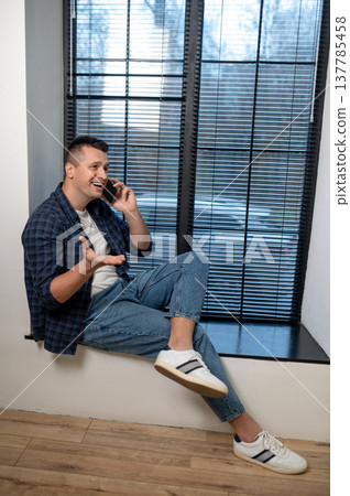 Laughing man sitting by window talking on smartphone in contemporary office Laughing man sitting by window talking on smartphone in contemporary office 137785458