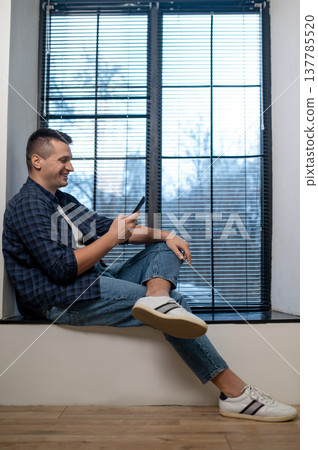 Cheerful man browsing smartphone on windowsill in modern office Cheerful man browsing smartphone on windowsill in modern office 137785520