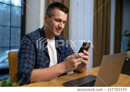 Happy man using smartphone in modern office interior with laptop Happy man using smartphone in modern office interior with laptop 137785550