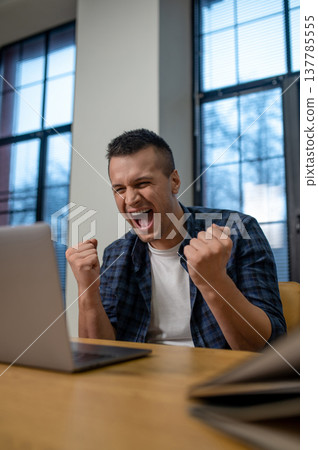 Excited man celebrating success with raised fists sitting at desk in modern office interior Excited man celebrating success with raised fists sitting at desk in modern office interior 137785555