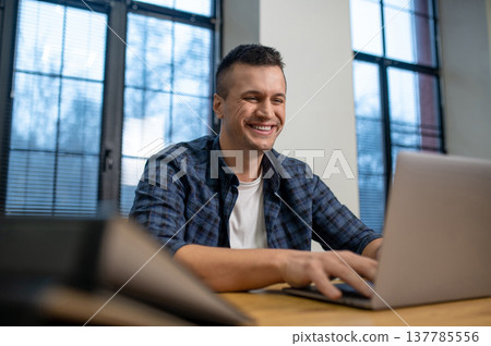 Cheerful man using laptop sitting in modern office with large windows Cheerful man using laptop sitting in modern office with large windows 137785556