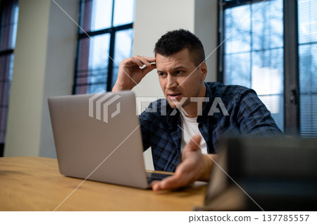 Concentrated man touching forehead while working on laptop at wooden table in office Concentrated man touching forehead while working on laptop at wooden table in office 137785557