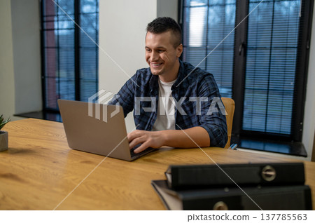 Smiling man using laptop sitting at wooden table in office interior with windows Smiling man using laptop sitting at wooden table in office interior with windows 137785563