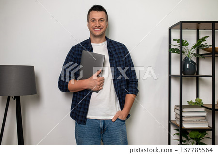 Positive man standing with laptop in home interior with shelf and lamp Positive man standing with laptop in home interior with shelf and lamp 137785564