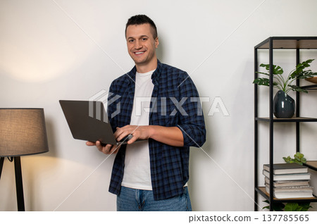 Smiling man holding laptop in home setting with shelf and lamp Smiling man holding laptop in home setting with shelf and lamp 137785565