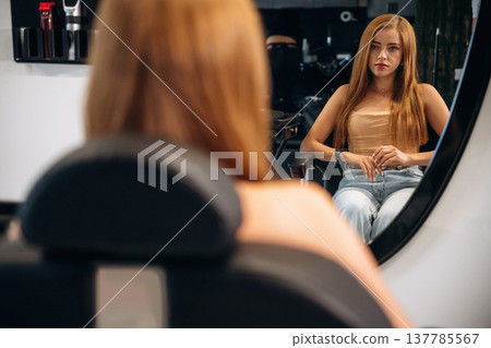Woman is in a beauty salon, sitting and waiting for stylist, looking at the mirror Woman is in a beauty salon, sitting and waiting for stylist, looking at the mirror 137785567