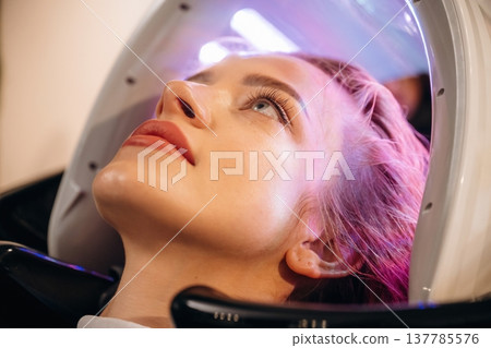 Pink or velvet color. Woman is in a beauty salon with LED hair growth helmet Pink or velvet color. Woman is in a beauty salon with LED hair growth helmet 137785576