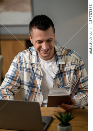 Positive man studying book while working on laptop at wooden desk Positive man studying book while working on laptop at wooden desk 137785588