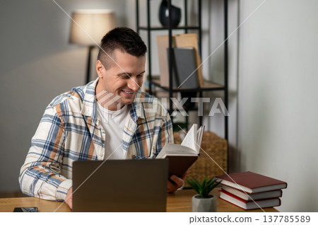 Cheerful man reading book and working on laptop in modern home workspace Cheerful man reading book and working on laptop in modern home workspace 137785589
