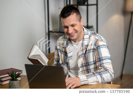 Smiling man multitasking with book and laptop in home office Smiling man multitasking with book and laptop in home office 137785590