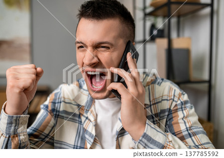Thrilled man celebrating success while talking on smartphone in home office interior Thrilled man celebrating success while talking on smartphone in home office interior 137785592