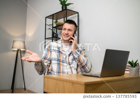 Excited man talking on smartphone and gesturing while sitting at wooden table Excited man talking on smartphone and gesturing while sitting at wooden table 137785593