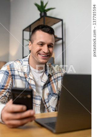 Cheerful man multitasking with laptop and smartphone at wooden table in home workspace Cheerful man multitasking with laptop and smartphone at wooden table in home workspace 137785594