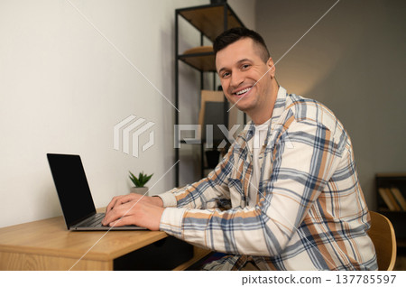 Happy man working on laptop at wooden desk in home office Happy man working on laptop at wooden desk in home office 137785597
