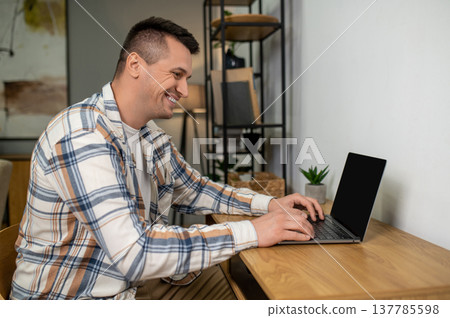 Cheerful man using laptop while sitting at wooden table in home office interior Cheerful man using laptop while sitting at wooden table in home office interior 137785598