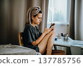 Teen girl sitting at home by the window with a smartphone, earphones and a bath towel on her head. Natural home lifestyle scene about messaging, music, privacy and everyday life. 137785756