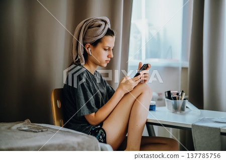 Teen girl sitting at home by the window with a smartphone, earphones and a bath towel on her head. Natural home lifestyle scene about messaging, music, privacy and everyday life. Teen girl sitting at home by the window with a smartphone, earphones and a bath towel on her head. Natural home lifestyle scene about messaging, music, privacy and everyday life. 137785756