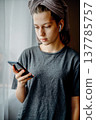 Teen girl standing at home by the window with a smartphone in her hand, earphones and a towel on her head. Natural home lifestyle scene about communication, music and a quiet morning moment. 137785757