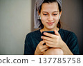 Home portrait of a teen girl with a slight smile and a smartphone in her hands. Earphones and a towel on her head support themes of a pleasant message, communication, music and positive emotion. 137785758