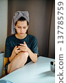 Teen girl sitting at a table at home and looking at her phone. The towel on her head and earphones create a story about music, communication, study and quiet daily life. 137785759
