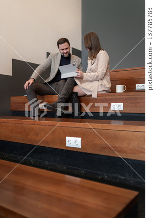 Two coworkers reviewing notes and discussing project on wooden benches in a neutral office setting. Review, notes, project, office, teamwork. 137785783