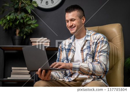 Smiling man using laptop while sitting in office chair in modern interior Smiling man using laptop while sitting in office chair in modern interior 137785804