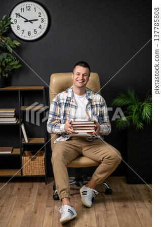 Joyful man holding stack of books sitting in office chair in interior with wall clock Joyful man holding stack of books sitting in office chair in interior with wall clock 137785808