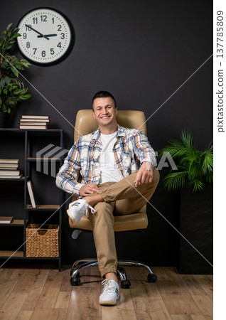 Happy man sitting in office chair posing in interior with wall clock and bookshelf Happy man sitting in office chair posing in interior with wall clock and bookshelf 137785809