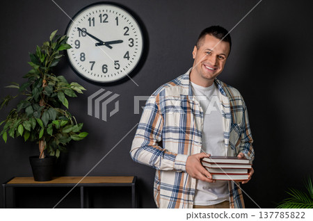 Smiling man holding stack of books standing in modern office interior Smiling man holding stack of books standing in modern office interior 137785822