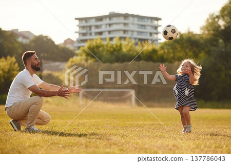 Throwing soccer ball high up. Father with his little daughter are on the field Throwing soccer ball high up. Father with his little daughter are on the field 137786043