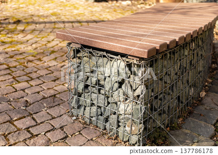 A bench is seen with a wooden top and a base made of stones held together by wire. It sits on a path made of cobblestones. The setting is bright and clear A bench is seen with a wooden top and a base made of stones held together by wire. It sits on a path made of cobblestones. The setting is bright and clear 137786178